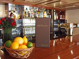 Elegante Schiffsbar auf der MS Florentina mit polierter Holztheke, Obstkorb mit Orangen und Limetten, Blumenarrangement und gut sortierter Bar.