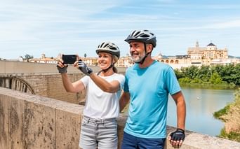 Zwei Radfahrer mit Helmen machen ein Selfie auf einer Steinbrücke in Andalusien. Historische Gebäude und ein Fluss sind im Hintergrund sichtbar.