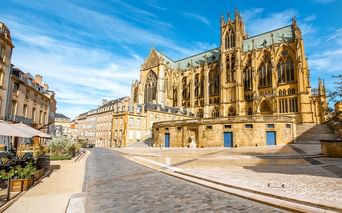 Gotische Kathedrale mit verzierten Türmen in Metz, Frankreich. Das gelbe Steingebäude überblickt einen gepflasterten Platz mit Wohngebäuden links.