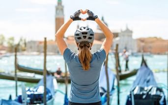 Radfahrerin mit Helm formt Herz mit Händen vor Venedigs Lagune mit Gondeln und dem Campanile von San Giorgio Maggiore.