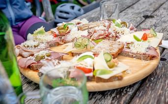 Holzbrett mit belegten Broten mit Schinken, Käse, Gurke und Gemüse auf Picknicktisch. Fahrradausrüstung und Helme sichtbar.
