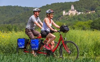 Couple on red tandem bike with blue panniers cycling through yellow flowering field in Altmühltal Nature Park, castle on forested hill in background.