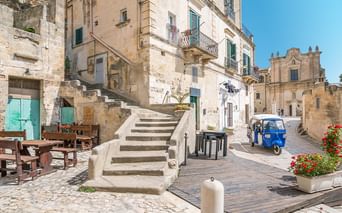 Kopfsteinpflasterplatz in Apulien mit Steintreppe, verwitterten Gebäuden, blauer Ape und barocker Kirche unter blauem Himmel.