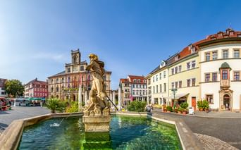 Prunkvoller Barockbrunnen mit goldener Statue in der Mitte eines Kopfsteinpflaster-Marktplatzes umgeben von bunten historischen Gebäuden.