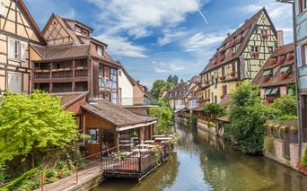 Schmaler Kanal zwischen bunten Fachwerkhäusern im Süd-Elsass. Holzterrasse ragt über das Wasser, üppiges Grün säumt die Ufer.