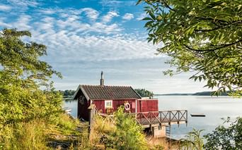 Rotes Holz-Bootshaus mit Wellblechdach und Holzsteg, der sich in ruhiges Seewasser erstreckt, umgeben von grünen Bäumen unter bewölktem Himmel.