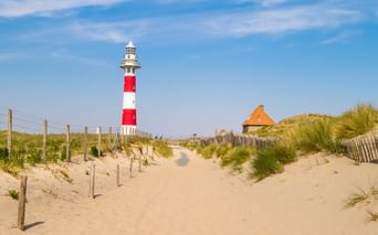 Sandweg durch Dünen führt zu rot-weiß gestreiftem Leuchtturm unter blauem Himmel. Holzzäune säumen den Weg, mit Strandgras und einem Gebäude.