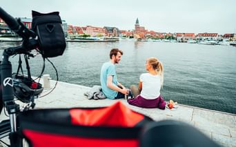 Zwei Radfahrer sitzen mit ihren Rädern am Wasser und blicken auf den Hafen und die historische Stadt mit roten Dächern und Kirchturm.