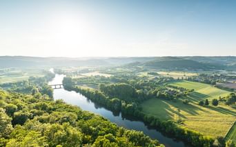 Luftaufnahme eines gewundenen Flusses durch grüne Felder und Wälder im Périgord. Hügel und eine Brücke sind unter klarem Himmel sichtbar.