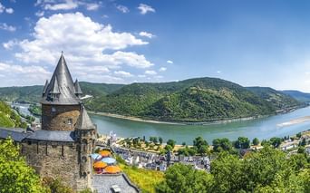 Stone castle tower overlooking Rhine River bend with green hills and village. Blue sky with white clouds above the scenic river valley.