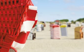 Nahaufnahme eines rot-weiß gestreiften Strandkorbs am Sandstrand in Friesland, mit Reihen bunter Strandkörbe im Hintergrund.