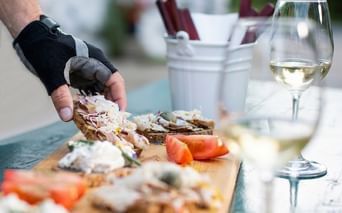Hand with cycling glove holding open sandwich on wooden board. White wine glass and cutlery in background. Fresh tomatoes visible.