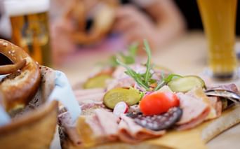 Wooden board with Bavarian snacks: pretzels, cold cuts, pickles, cherry tomato, and rosemary. Beer glasses visible in background.