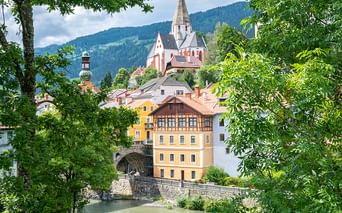 Alpenstadt mit Kirchturm und gelbem Gebäude über Fluss, eingerahmt von grünen Bäumen. Bewaldete Berge im Hintergrund unter bewölktem Himmel.