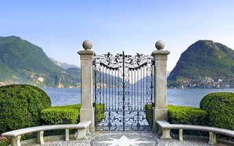 Verziertes schmiedeeisernes Tor mit Steinpfeilern, flankiert von geschwungenen Bänken und gepflegten Hecken, mit Blick auf Lago Maggiore.