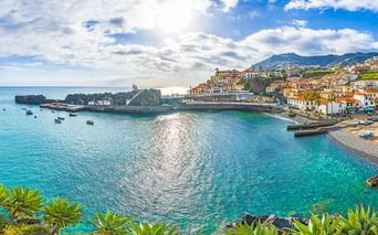 Küstenort auf Madeira mit weißen Gebäuden und roten Dächern über einer türkisen Bucht. Berge erheben sich im Hintergrund unter blauem Himmel.