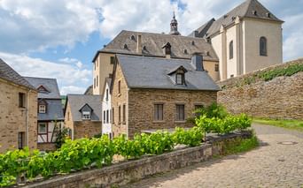 Kopfsteinpflasterstraße mit Weinreben, die zu traditionellen Steinhäusern und einer Kirche mit Turm an der Mosel führt, unter bewölktem Himmel.