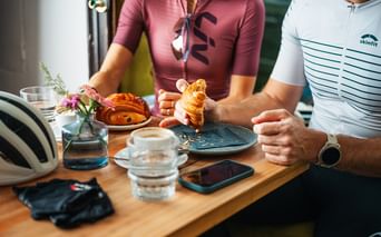 Zwei Radfahrer in Radtrikots an einem Cafétisch mit Croissants und Kaffee. Ein Helm und Handschuhe liegen auf dem Holztisch.