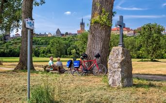 Zwei Radfahrer sitzen auf einer Bank unter einem großen Baum mit roten Tourenrädern daneben. Ein Steinkreuz und Radwegeschild sind sichtbar, mit historischer Stadtsilhouette im Hintergrund.