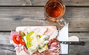 Wooden board with sliced ham, bacon, cheese, tomato, cucumber, and peppers beside a glass of amber tea on a weathered wooden table.