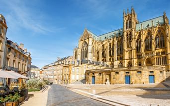 Gotische Kathedrale in Metz mit verzierten Türmen und Strebepfeilern, umgeben von historischen gelben Steingebäuden und einem Kopfsteinpflasterplatz.