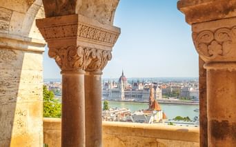 View through ornate stone arches with carved capitals overlooking the Danube River and Hungarian Parliament building in Budapest.