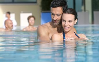 Lächelndes Paar im Hallenbad mit anderen Gästen im Hintergrund. Moderne Wellnessanlage mit Glasfenstern und sichtbaren Pflanzen.