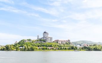 Die Esztergom Basilika mit ihrer markanten Kuppel krönt einen grünen Hügel über der Donau, umgeben von Bäumen und Gebäuden unter blauem Himmel.
