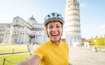 Lächelnde Radfahrerin in gelbem Trikot und Helm macht Selfie auf der Piazza dei Miracoli mit Schiefem Turm von Pisa und Dom im Hintergrund.
