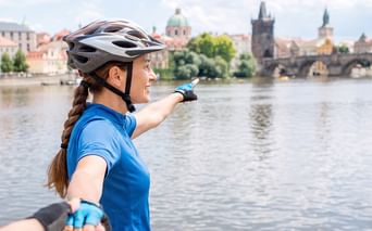 Radfahrerin in blauem Shirt und Helm zeigt auf die Karlsbrücke und historische Prager Stadtlandschaft über die Moldau.