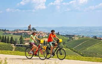 Zwei Radfahrer fahren durch hügelige Weinberge mit einem historischen italienischen Dorf im Hintergrund unter blauem Himmel mit weißen Wolken.