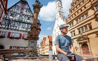Radfahrer mit Helm rastet am Brunnen auf historischem Marktplatz. Fachwerkhäuser, verzierte Säule und weißer Kirchturm bei blauem Himmel.