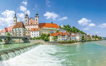 Historische Stadt am Flussufer mit barocken Kirchtürmen und bunten Gebäuden entlang der Enns. Ein Wehr erzeugt Wildwasser im Vordergrund.