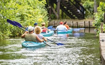 Gruppe von Paddlern in türkisfarbenen Kajaks auf einem schmalen Kanal im Spreewald, umgeben von üppiger grüner Vegetation und einer Holzbrücke.