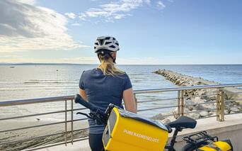 Radfahrerin mit weißem Helm und gelben Taschen pausiert an Küstenpromenade in Slowenien mit Blick auf ruhiges blaues Meer und Steinmole.
