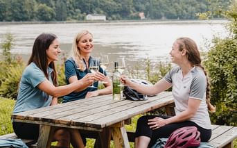 Drei Radfahrerinnen stoßen mit Weißwein an einem Holztisch an, mit Blick auf einen See in Oberösterreich und bewaldeten Hügeln im Hintergrund.