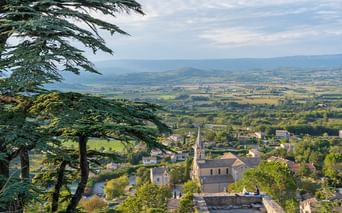Elevated view of a Provençal village with a church and stone buildings, framed by cedar trees. Rolling agricultural valley extends to distant mountains.