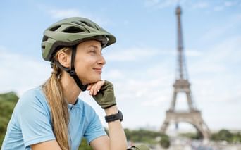 Radfahrerin mit grünem Helm und blauem Shirt, im Hintergrund verschwommen der Eiffelturm unter blauem Himmel in Paris.