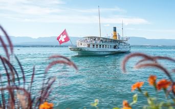 Historic paddle steamer with Swiss flag sailing on turquoise Lake Geneva. Orange flowers frame the foreground, mountains visible in background.