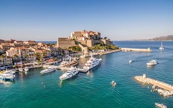 Luftaufnahme des Hafens von Calvi auf Korsika mit türkisfarbenem Wasser, Luxusyachten, historischer Zitadelle auf dem Hügel und Küstenstadt.