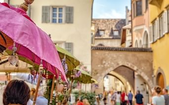 Pink decorative umbrella in foreground of a market street with historic archway and colorful buildings in an Italian town square.