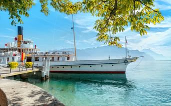 Weißes historisches Dampfschiff am Steinpier des Genfersees mit klarem türkisfarbenem Wasser, Bergen im Hintergrund und Ästen darüber.