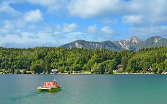 Tretboot mit gestreiftem Dach auf türkisfarbenem See in Kärnten, mit bewaldeten Ufern und Bergkette unter blauem Himmel mit Wolken.