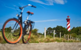 Fahrrad mit orangem Rad im Vordergrund auf sandigem Weg, rot-weiß gestreifter Leuchtturm hinter grüner Vegetation unter blauem Himmel in Zeeland.