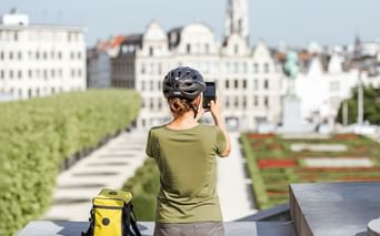 Radfahrerin mit Helm fotografiert Brüsseler Stadtbild mit historischen Gebäuden und formalen Gärten. Gelbe Fahrradtasche neben ihr.