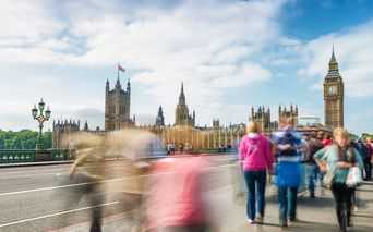 Motion-blurred pedestrians crossing Westminster Bridge in London with Big Ben and the Houses of Parliament visible under a cloudy sky.