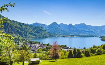 Green meadow overlooking a lake in Salzkammergut with a town on the shore and mountain range in the background under clear blue sky.