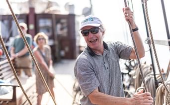 Mann mit weißer Kappe und Sonnenbrille lächelt auf Segelschiffsdeck und hält Takelage. Weitere Passagiere im Hintergrund an sonnigem Tag.