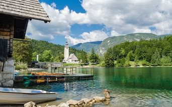 Smaragdgrüner Alpensee mit Bootshaus, bunten Booten und weißer Kirche mit Glockenturm. Berge und Wälder unter blauem Himmel mit Wolken.