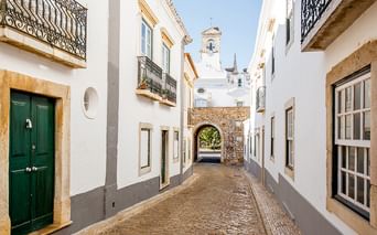 Enge Kopfsteinpflasterstraße in einem Algarve-Dorf mit weiß getünchten Häusern, Steinbogen und Kirchturm unter blauem Himmel.